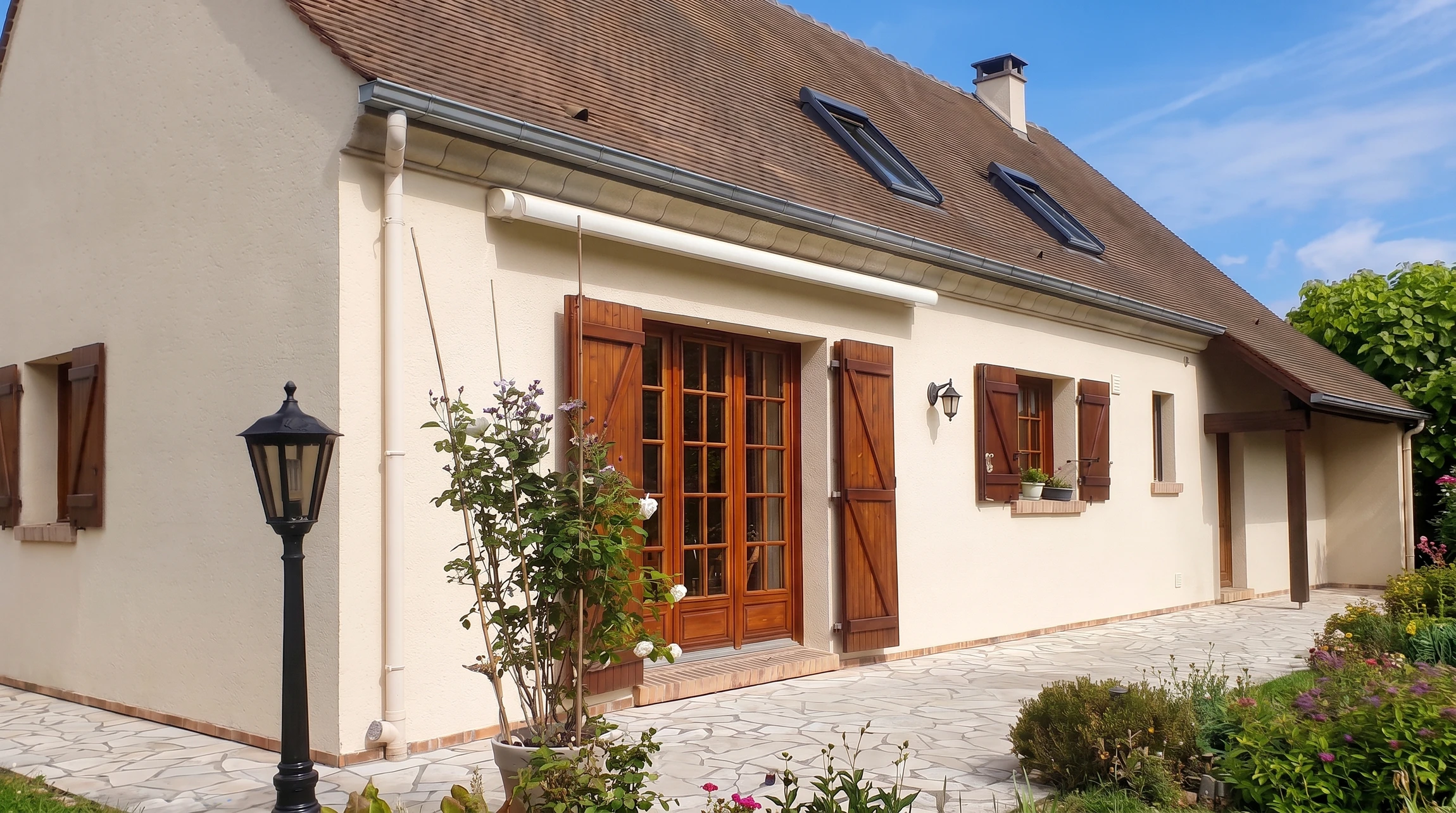 Maison pavillonnaire de plain-pied avec combles aménagés, façade enduit ton pierre, volets bois teinte chêne, porte-fenêtre à petits carreaux style français, lucarnes de toit type Velux, toiture tuiles mécaniques brun, gouttière PVC blanc, descente EP, applique murale, lampadaire fonte noir, terrasse dallage irrégulier, jardin fleuri