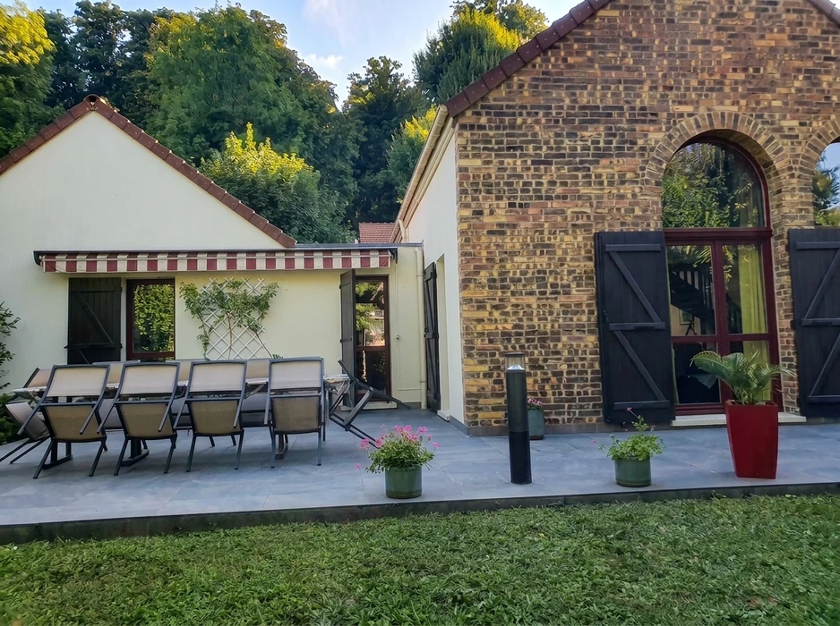 Terrasse extérieure pavée en carrelage grès cérame gris, attenante à une maison de caractère en briques apparentes avec fenêtre cintrée et volets bois foncé